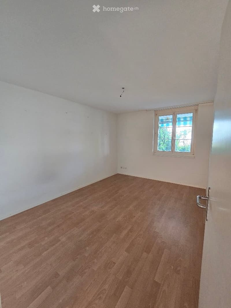 Empty room with wooden flooring and white walls, featuring a single window with white frame and a door with silver handle.