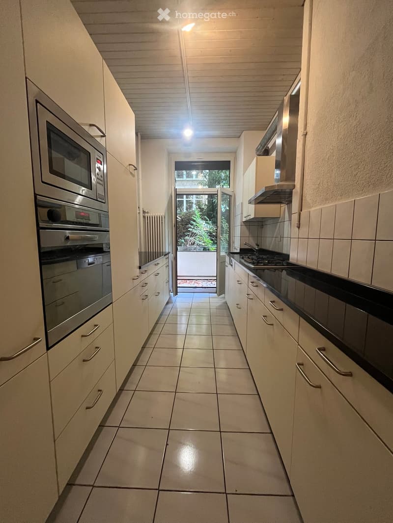 Modern kitchen with white cabinets, stainless steel appliances, tiled floor, and a view to the garden through an open doorway.