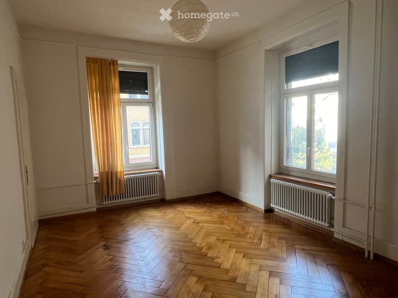 Empty room with wooden herringbone flooring, large windows with curtains, radiators, and a hanging light fixture.