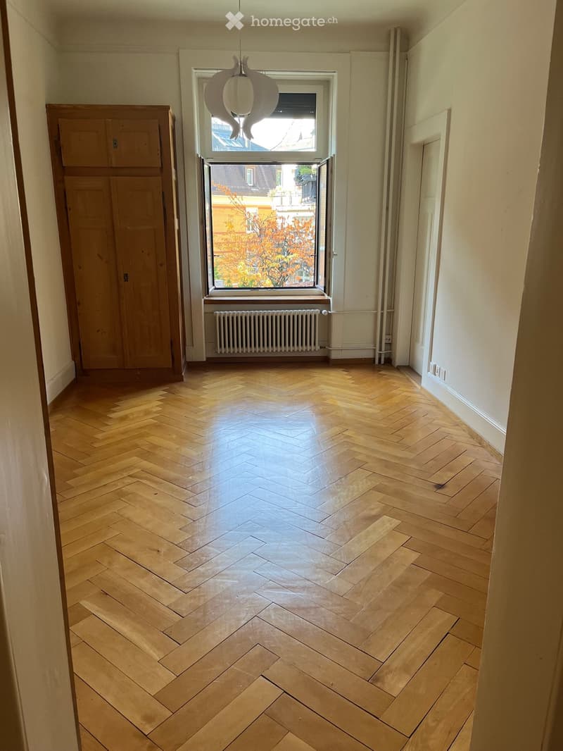 Empty hallway with herringbone wooden flooring, white walls, a window with a radiator below, and a wooden cabinet on the left side.