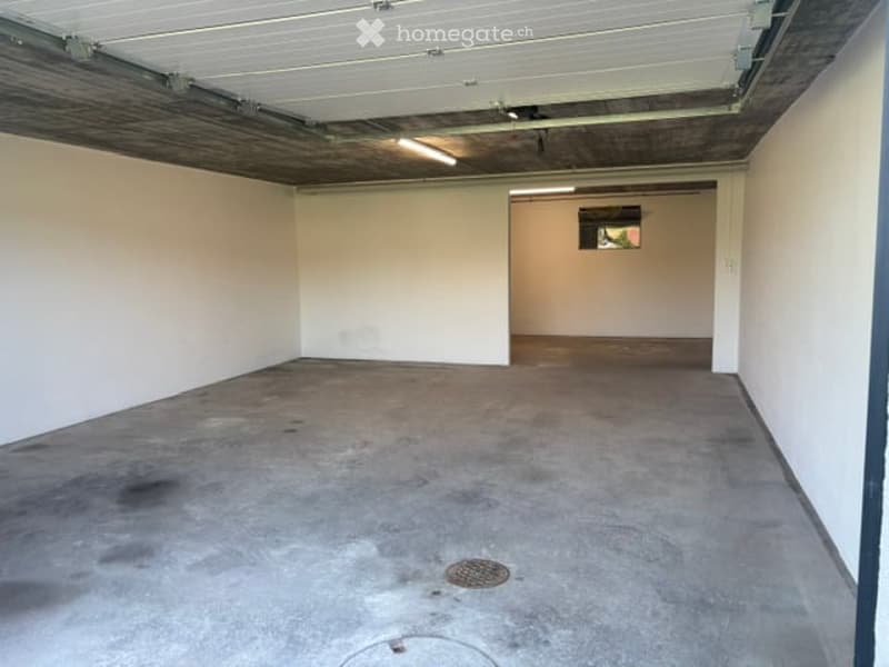 Empty garage with concrete floor, white walls, and a small adjacent room visible through an open doorway.