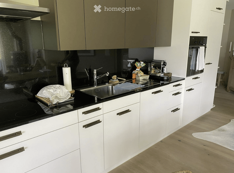 Modern kitchen with white cabinets, black countertops, stainless steel sink, and various appliances including a coffee machine and oven. Wooden flooring and a white rug are visible.