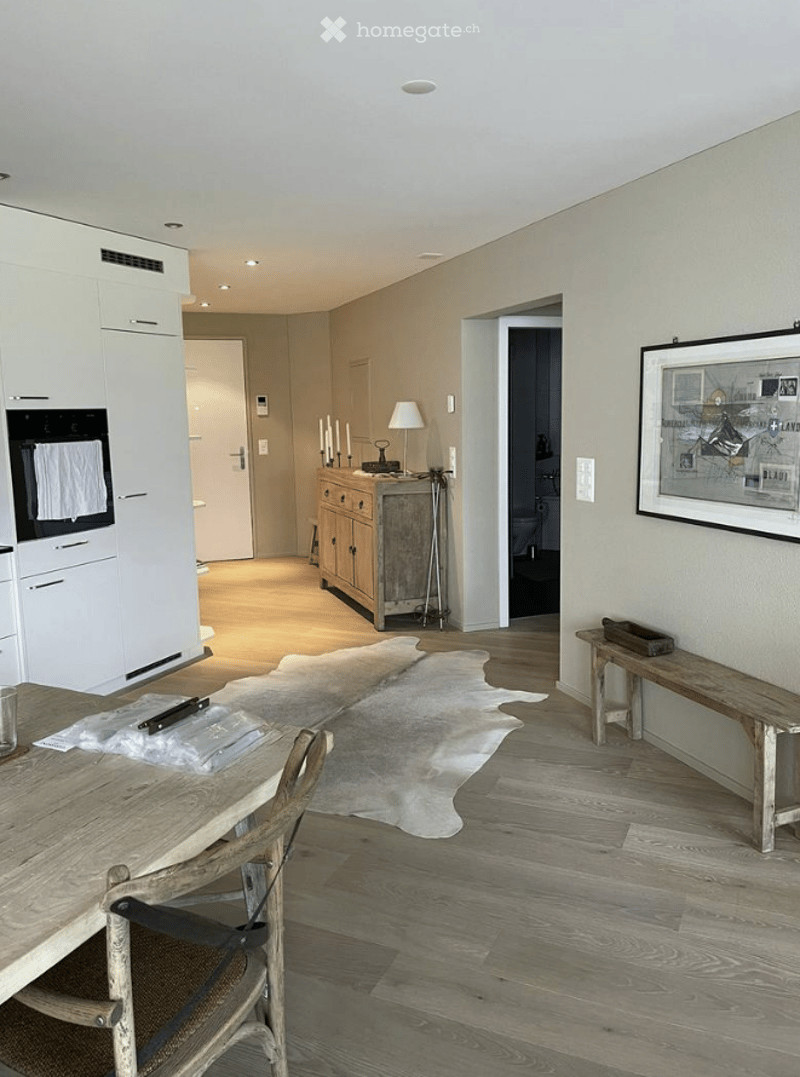 Bright, open-plan living and dining area with light wood flooring, white cabinetry, a wooden dining table with chairs, a bench, a small cabinet with a lamp, and a framed picture on the wall. A cowhide rug is placed on the floor.