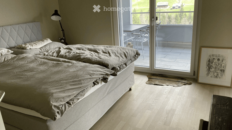 Modern bedroom with a large bed, grey bedding, a floor lamp, a picture frame, and a sliding glass door leading to a balcony with outdoor furniture.