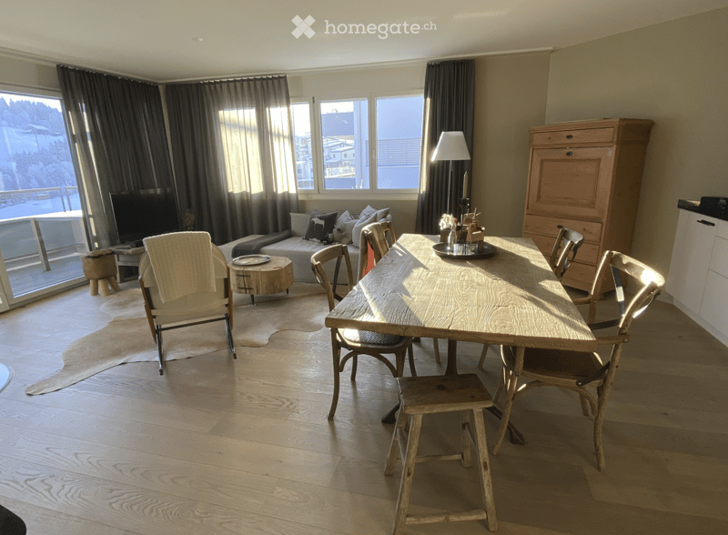 A well-lit living and dining area with wooden flooring, a large sofa, a dining table with chairs, a floor lamp, and a view through large windows.