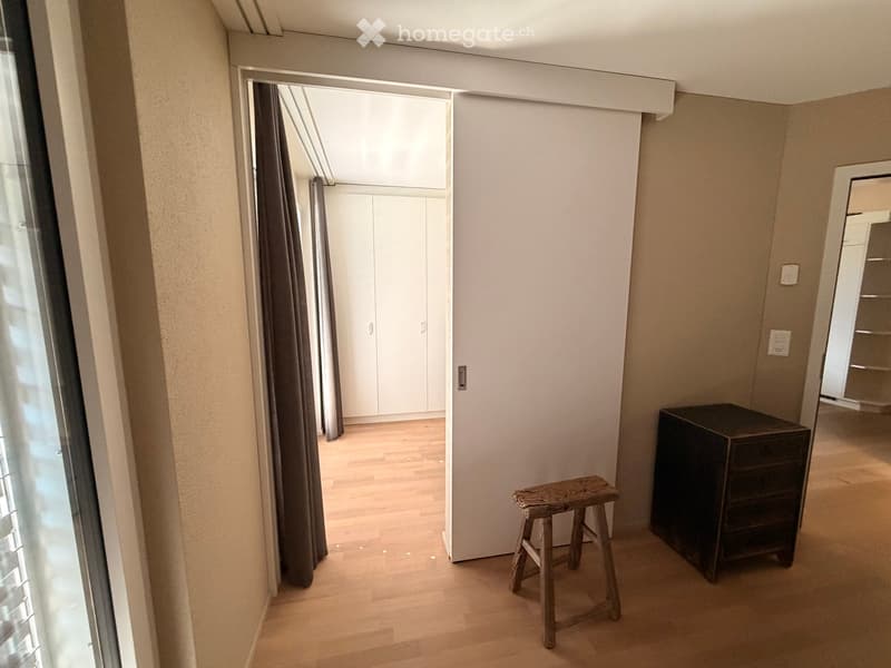 A small, well-lit room with wooden flooring, a wooden stool, a dark cabinet, and a closet with white doors visible through an open doorway.