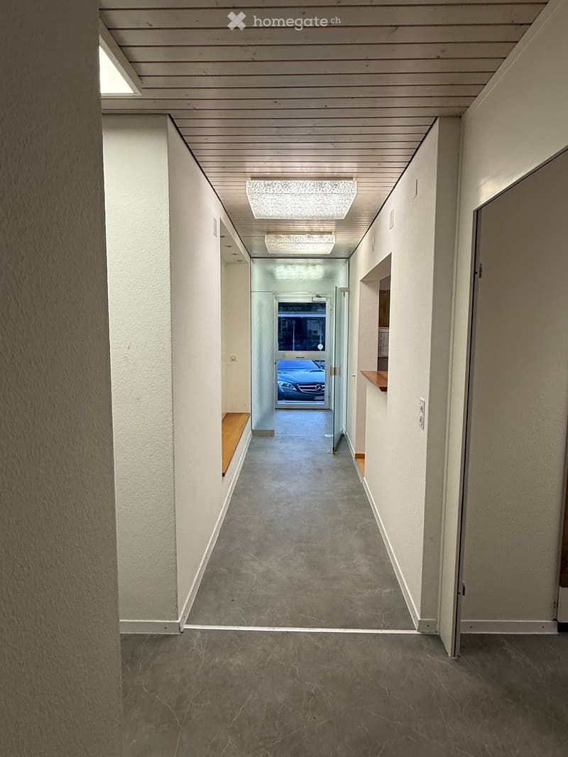 Modern hallway with white walls, grey carpeted floor, and recessed lighting. Features a bench on one side and a glass door leading to an outdoor area with a parked car visible.