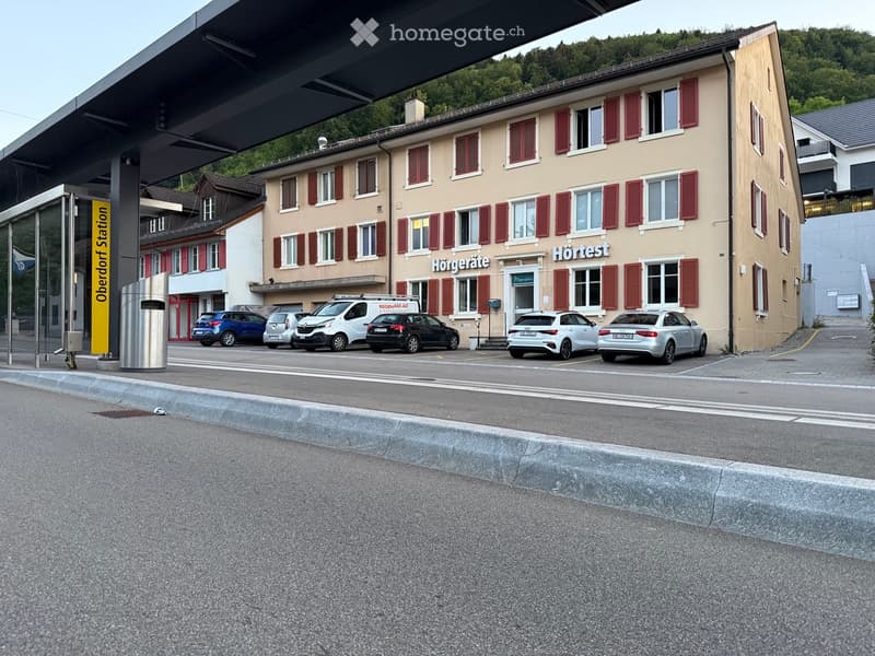 Two-story building with beige walls, red shutters, and multiple windows. Signs read "Hörgeräte" and "Hörtest". Several cars are parked in front, and a station sign is visible on the left.