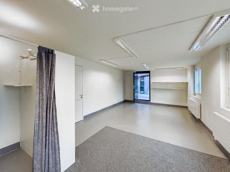 Empty room with white walls, grey carpet, and a curtain on the left side. The room has a door leading to another space and a window on the right side.