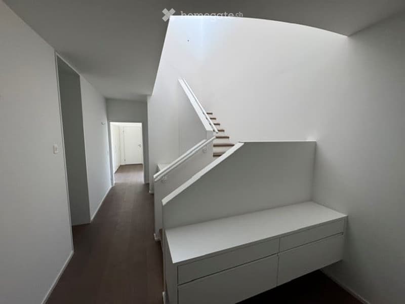 Modern hallway with white walls, wooden flooring, and a staircase leading upwards. The hallway features built-in storage units and a neutral color palette.