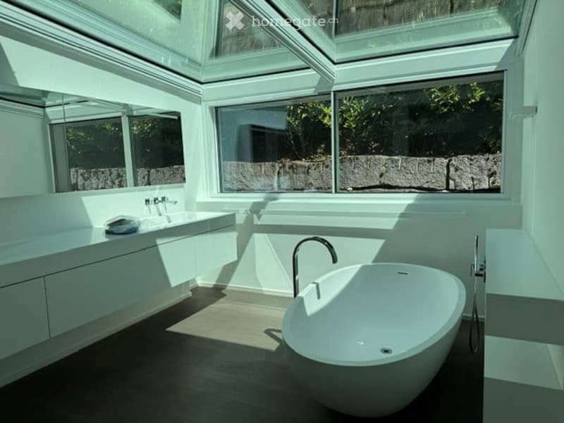 Modern bathroom with a large, freestanding bathtub, a sleek sink, and large windows offering natural light and a view of greenery outside.