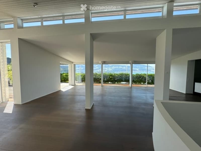 Bright, open-plan unfurnished room with wooden flooring, white walls, large windows offering a scenic view, and a balcony area visible.