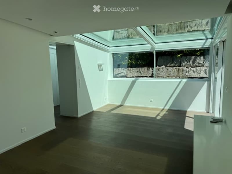 Completely empty room with wooden flooring, white walls, and large windows with a view of a stone wall outside. The room has a skylight and a small built-in bench along one wall.