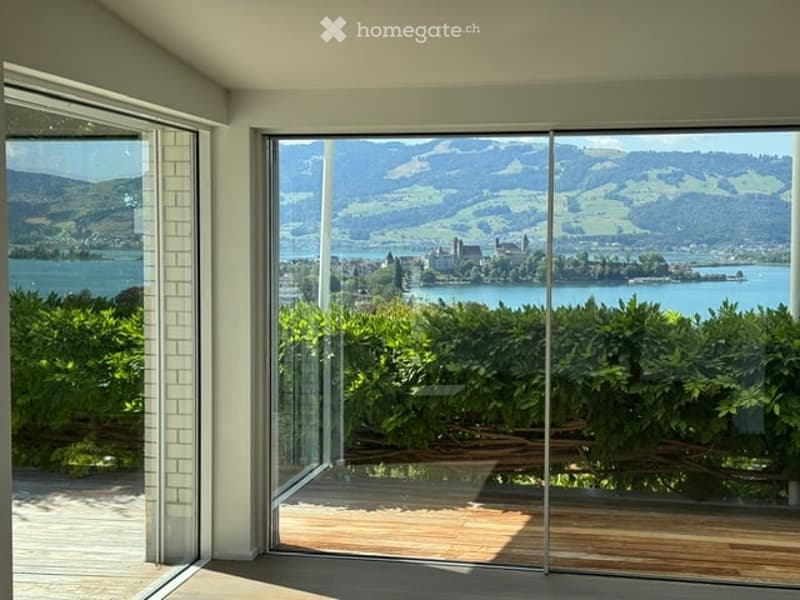 Large glass sliding doors open to a wooden deck, offering panoramic views of a lake and surrounding mountains.