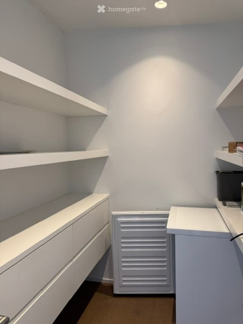 Empty room with white walls, built-in white shelving units, and a radiator. The floor is wooden, and the room is well-lit with recessed ceiling lights.