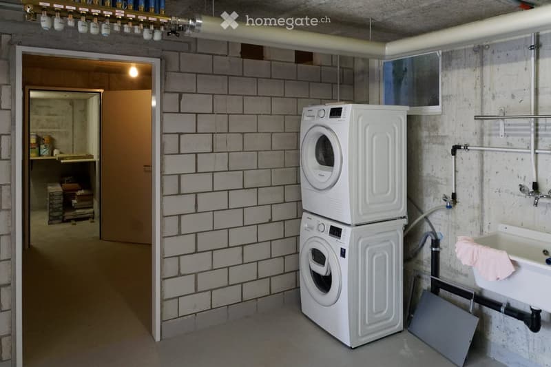 Utility room with white tiled walls, a stacked washing machine and dryer, a sink with a pink towel, and exposed pipes.