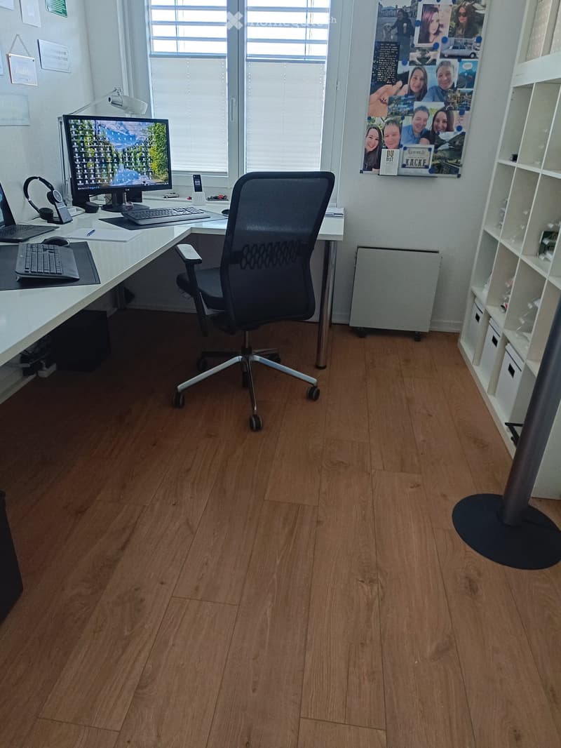 Office space with wooden flooring, large window, desk with computer setup, ergonomic chair, shelves, and personal decorations.