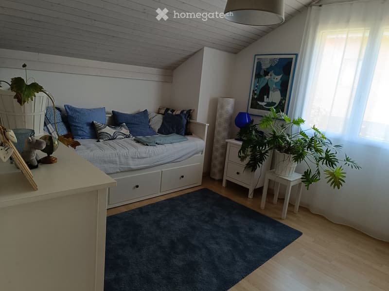 Cozy bedroom with a bed, nightstand, small table, and decorative plants. Features a blue rug, wooden floor, and a slanted ceiling.