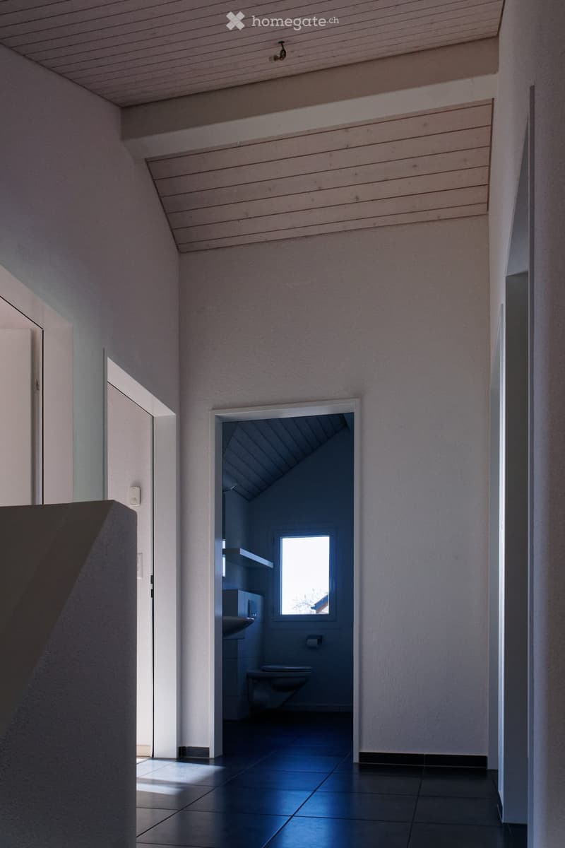 Modern hallway with sleek white walls, dark tiled floor, and an open doorway leading to a small bathroom with a window, sink, and toilet.