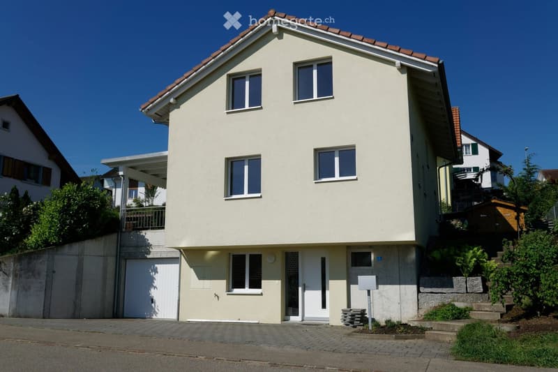 Two-story house with beige exterior, red-tiled roof, white-framed windows, and a garage on the lower level.