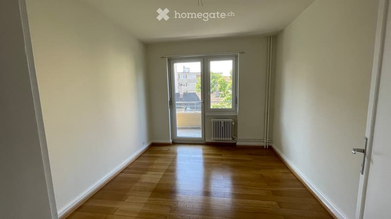 Empty room with wooden flooring, white walls, and a large window with a view of the outside. A radiator is visible under the window.