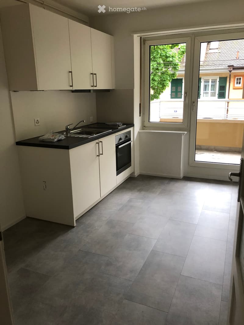 Modern kitchen with white cabinets, black countertop, sink, and oven. Large sliding glass door leads to a balcony with a view of greenery and neighboring buildings.