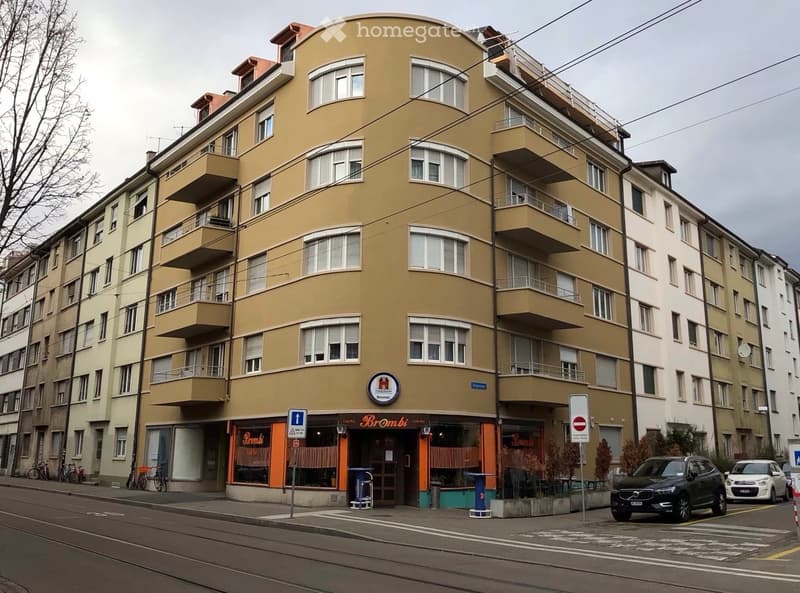 Multi-story residential building with balconies, storefront labeled "Brombi" at the ground level, and parked cars on the street.