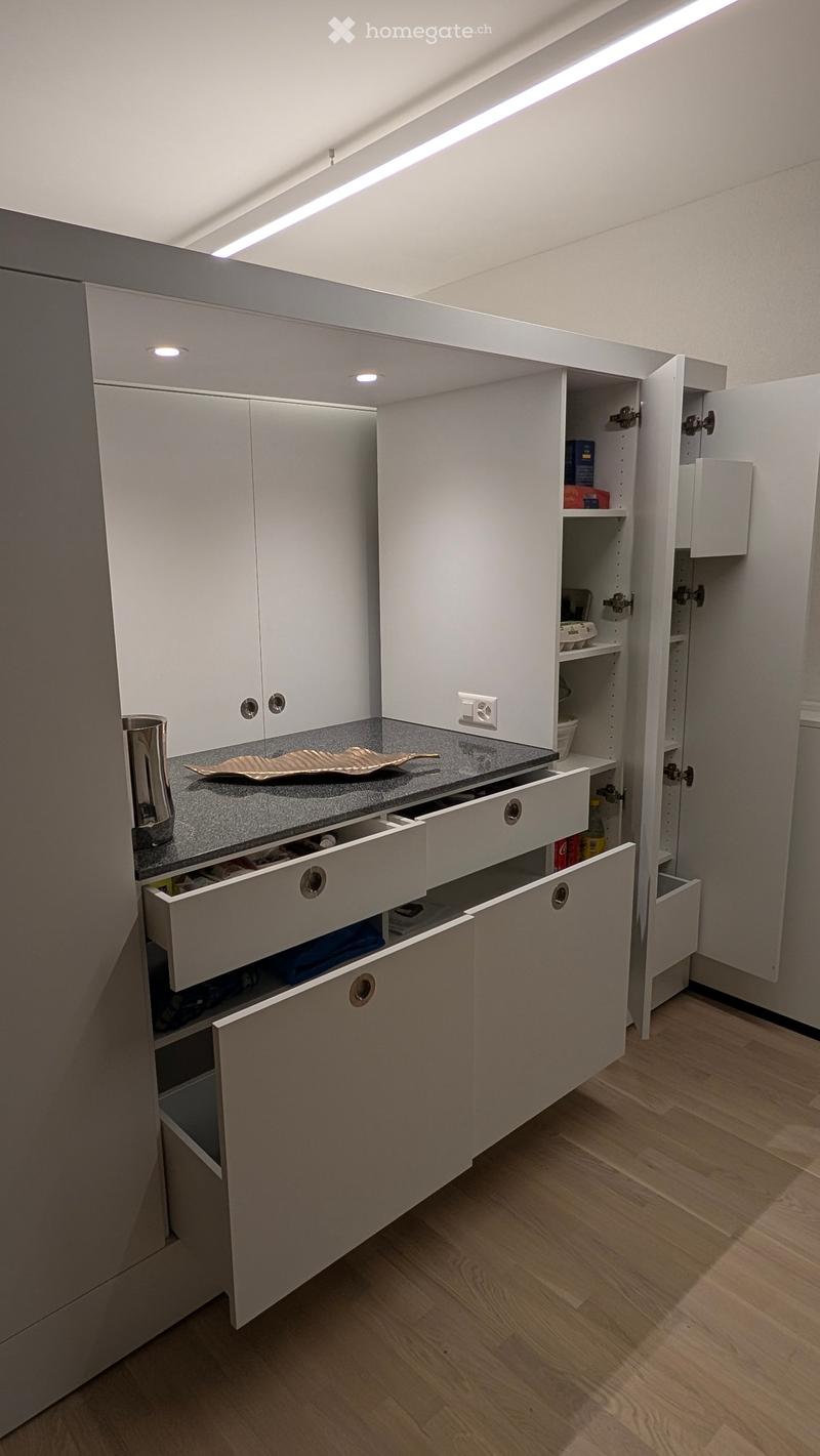 Modern kitchen with white cabinets, open shelving, and a grey countertop. Drawers and cabinets are visible, with a decorative plate and cup on the counter.