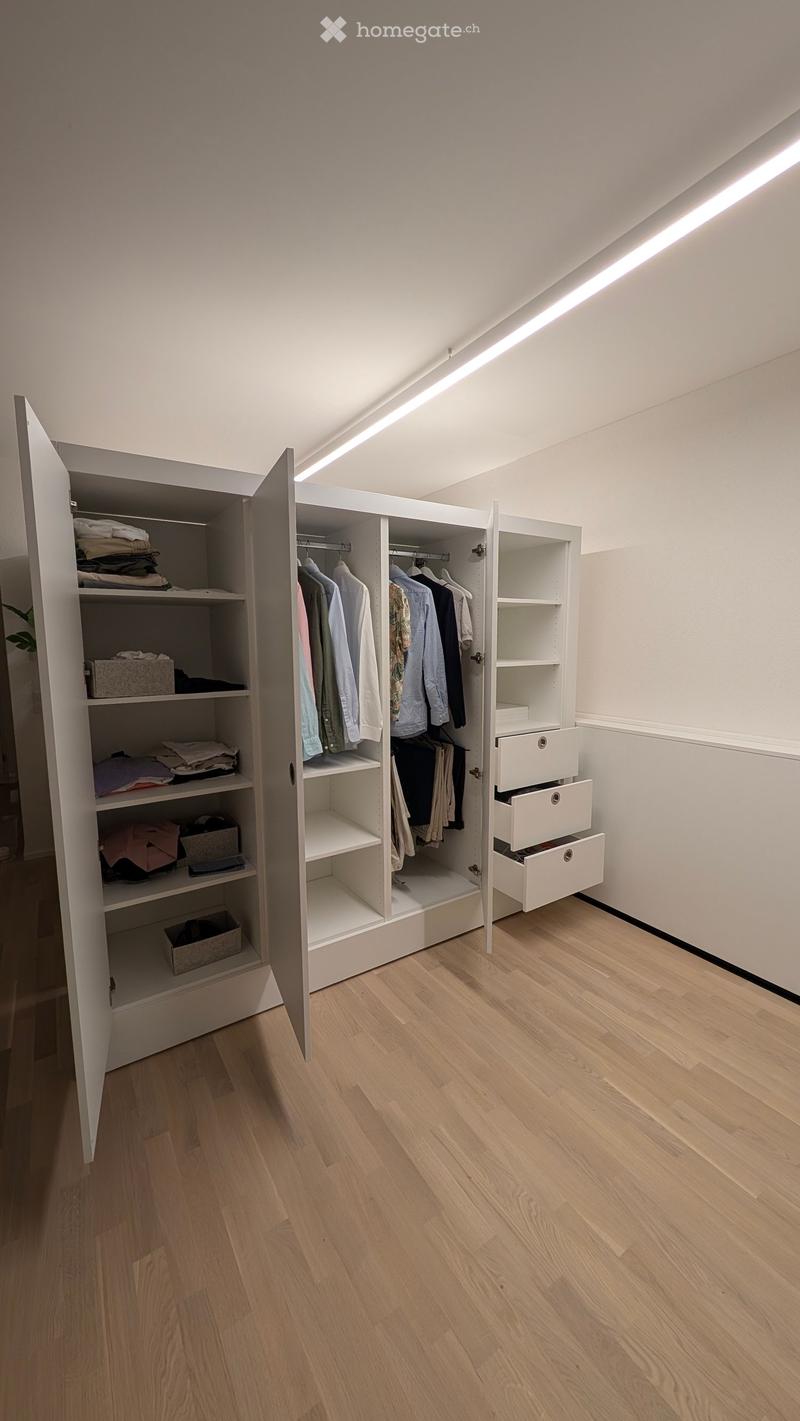 An empty room with light wooden flooring and white walls, featuring an open wardrobe with shelves and drawers, but no furniture or personal items present.
