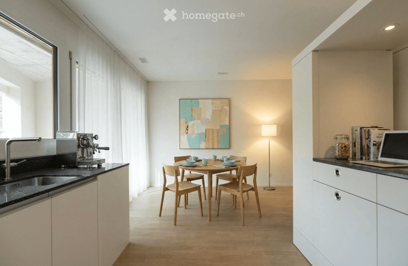 Modern kitchen and dining area with white cabinets, black countertops, a coffee machine, a round wooden dining table with four chairs, a laptop on the counter, and a floor lamp near the table.
