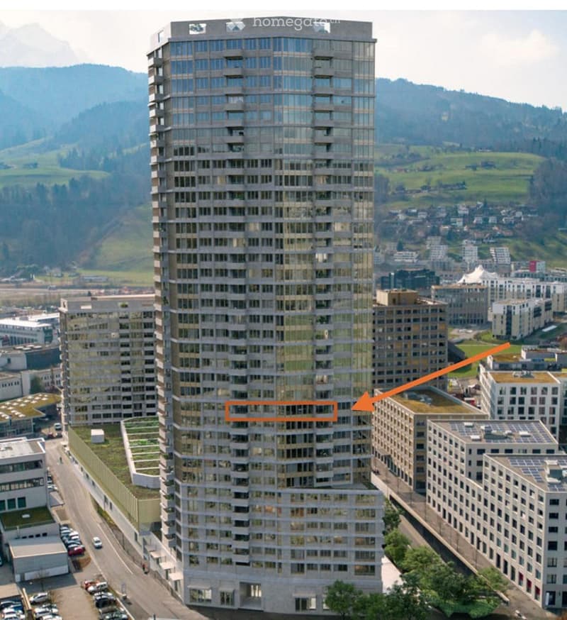 A modern high-rise building with a glass facade, surrounded by other structures and a parking lot.