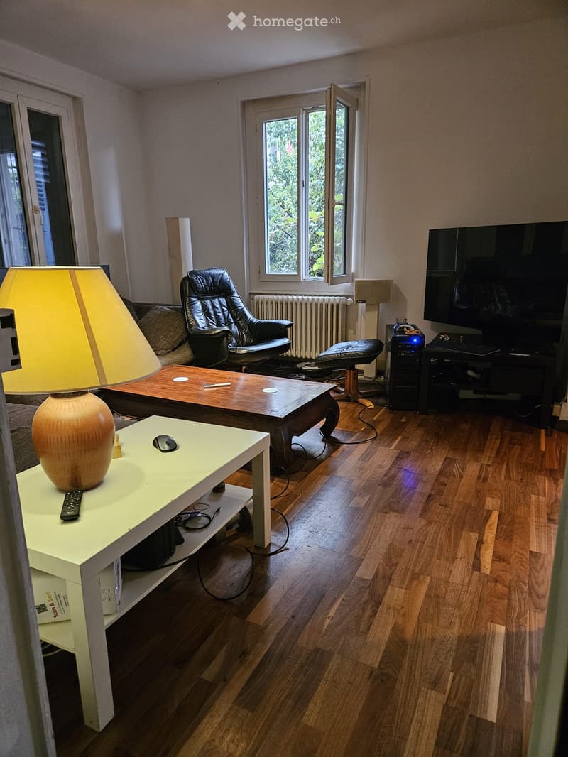 Living room with wooden flooring, large windows, sofa, coffee table, armchair, lamp, and computer setup.