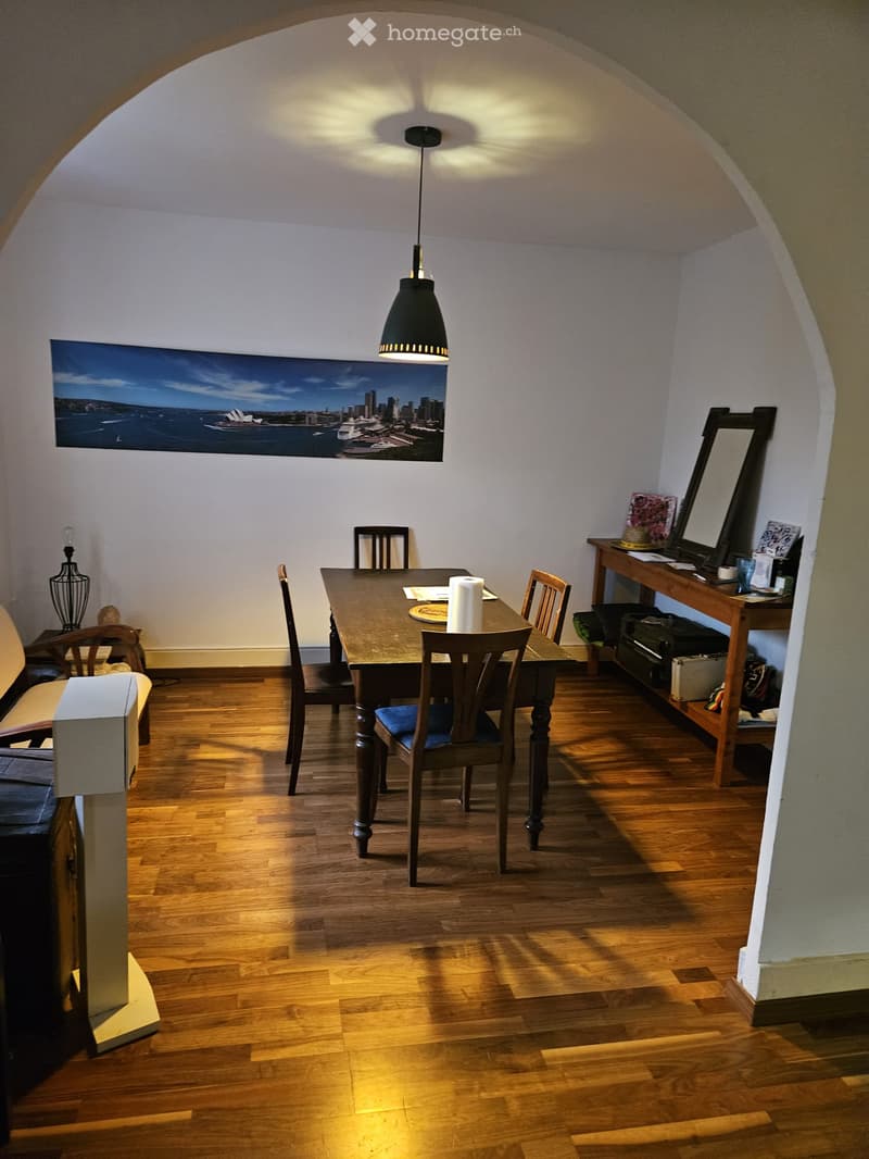 Dining area with wooden floor, dining table, chairs, sofa, and a mirror on a side table.
