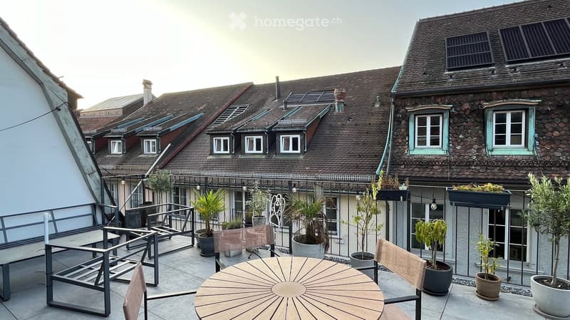 Courtyard with wooden table, chairs, potted plants, and metal railing, surrounded by historic buildings with tiled roofs and stone facades.