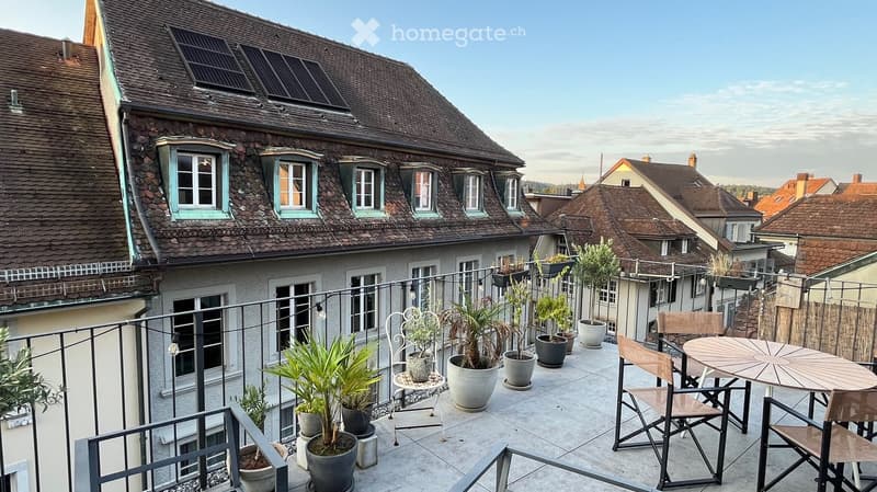 Balcony with a round table, chairs, and several potted plants, overlooking a cityscape with traditional houses and tiled roofs.