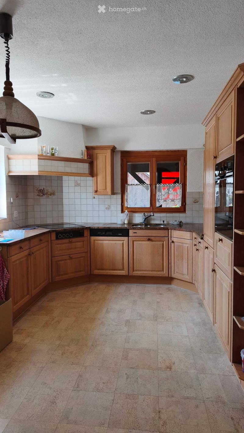 Bright kitchen with wooden cabinets, tiled backsplash, and a central island with built-in appliances including an oven and dishwasher. The space features a window with white curtains and a pendant light hanging from the ceiling.