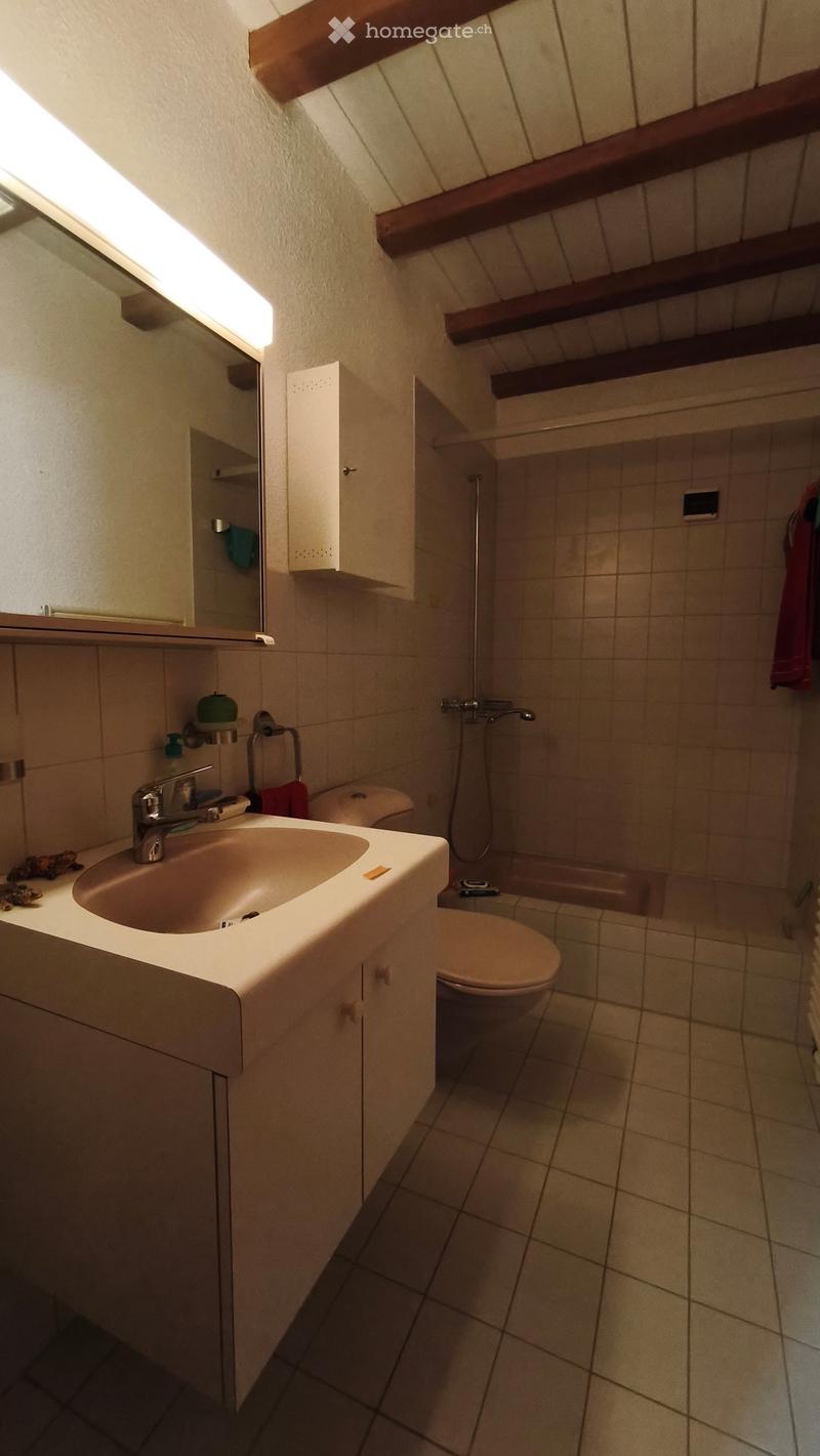 Bathroom with a sink, toilet, bathtub, mirror, and cabinet. The room features tiled walls and floor, with wooden beams on the ceiling.