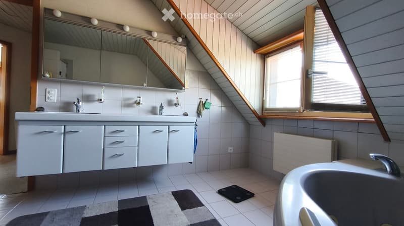 Modern bathroom with a double sink, large mirror, bathtub, and radiator. The room features white tiles, a window with blinds, and a patterned rug on the floor.