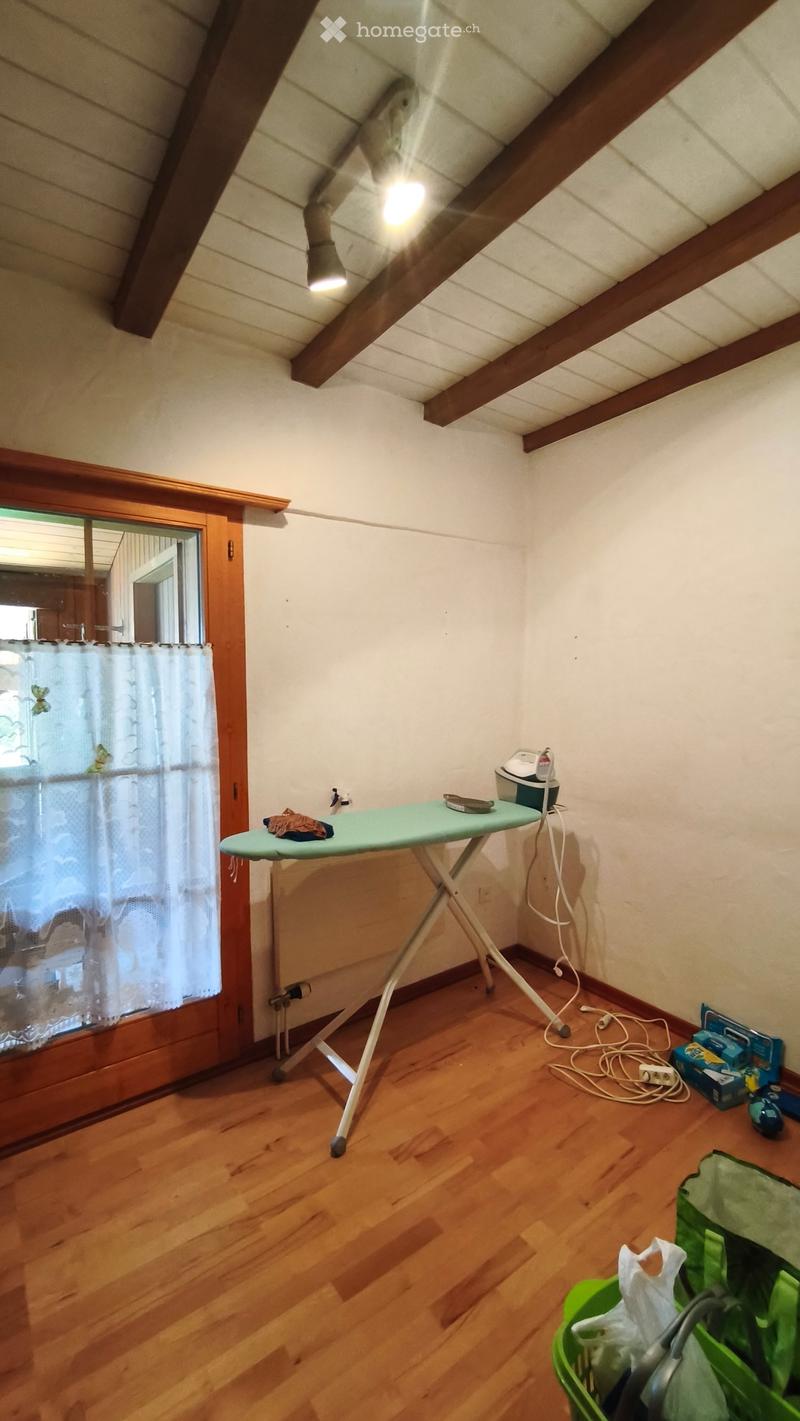 A small room with wooden flooring and white walls, featuring an ironing board with an iron placed on it, a basket, and a window with a curtain.