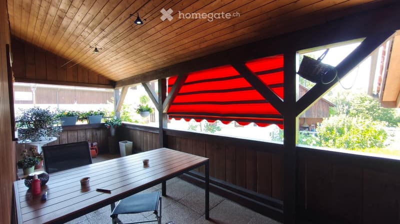 Wooden balcony with a table, chairs, and potted plants, featuring a red awning and a view of greenery.