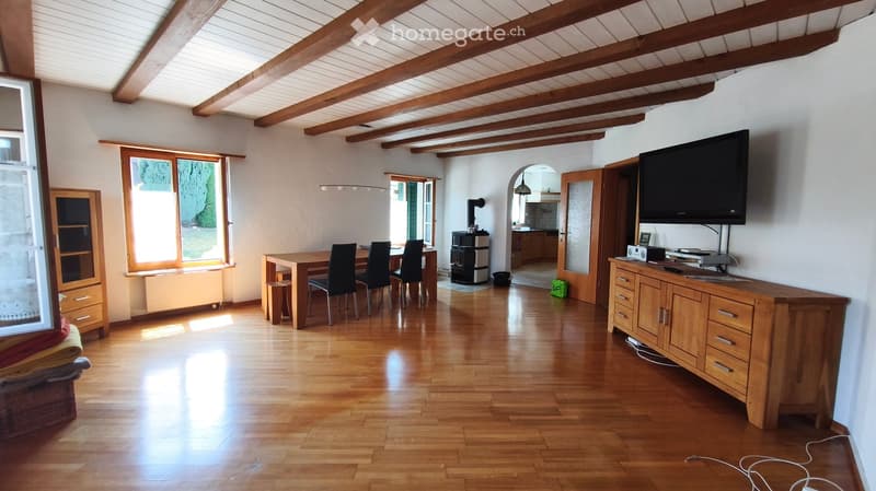 Bright living room with wooden floors, exposed wooden beams, large windows, a sofa, a dining table with chairs, a television on a wooden cabinet, and a radiator near the window.