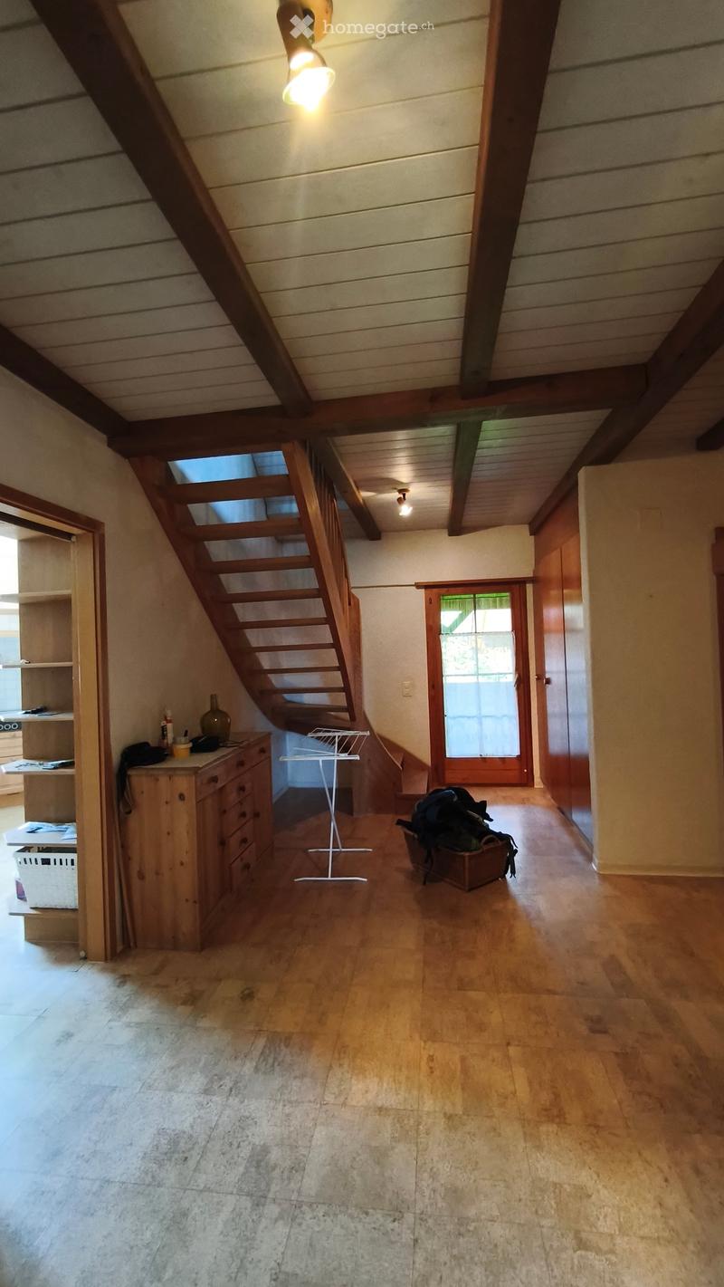 The image shows a spacious, well-lit hallway with wooden flooring and exposed wooden beams on the ceiling. A wooden staircase leads upwards, and a small wooden drawer unit is positioned near the staircase. A vase is placed on top of the drawer unit, and a