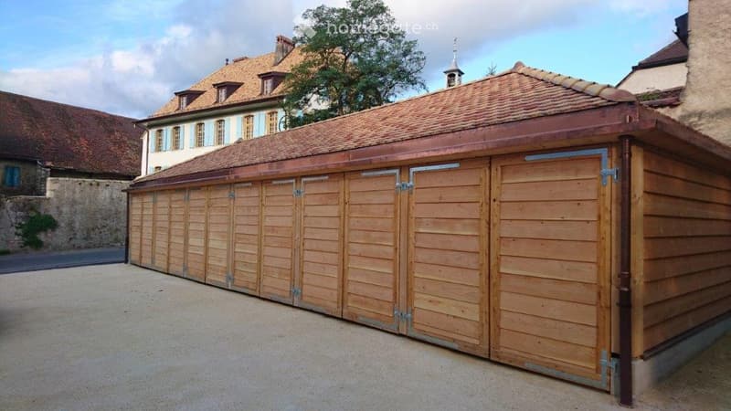 A large wooden garage with multiple doors, situated in a courtyard with traditional stone buildings and trees in the background.