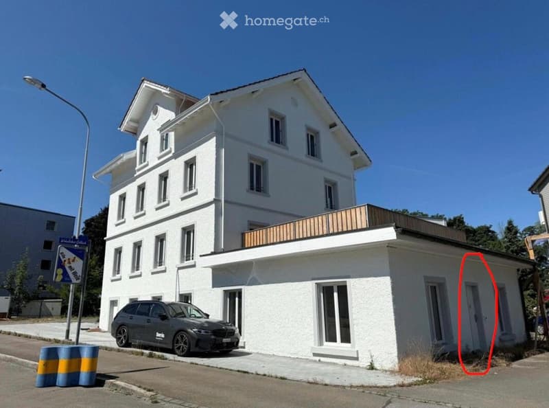 Two-story white building with multiple windows and a balcony on the roof. A car is parked in front.