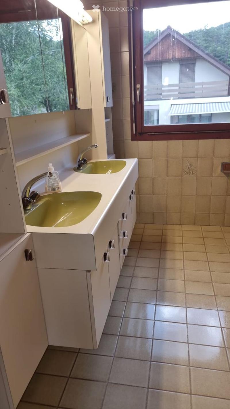 Bathroom with double sink, beige tiles, white cabinets, and a large window overlooking a house and greenery.