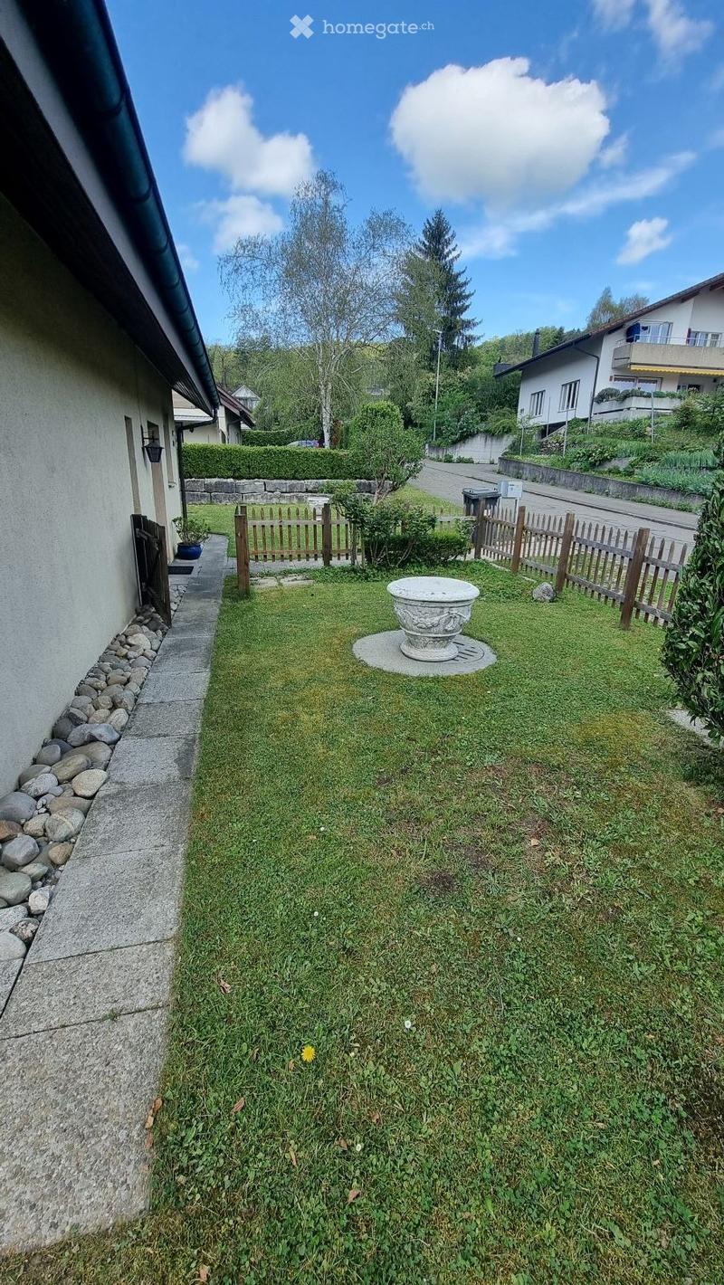 A small, well-maintained garden area with a stone pathway, a wooden fence, a decorative stone planter, and surrounding greenery.
