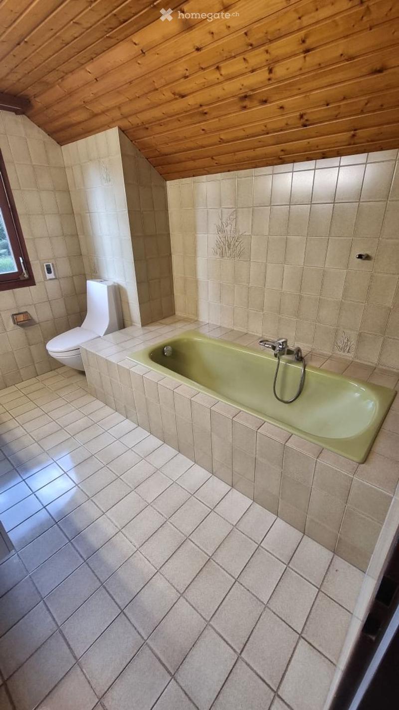 Bathroom with a green bathtub, toilet, tiled walls and floor, and a wooden slanted ceiling.