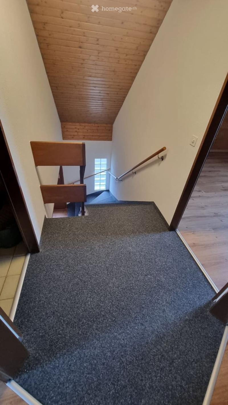 Carpeted staircase with wooden handrail and banister, leading to an upper level with wooden ceiling and white walls.