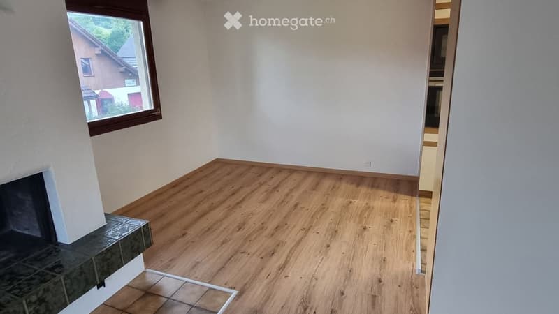 Empty room with wooden flooring, white walls, and a fireplace. There is a window showing a view of a neighboring house.