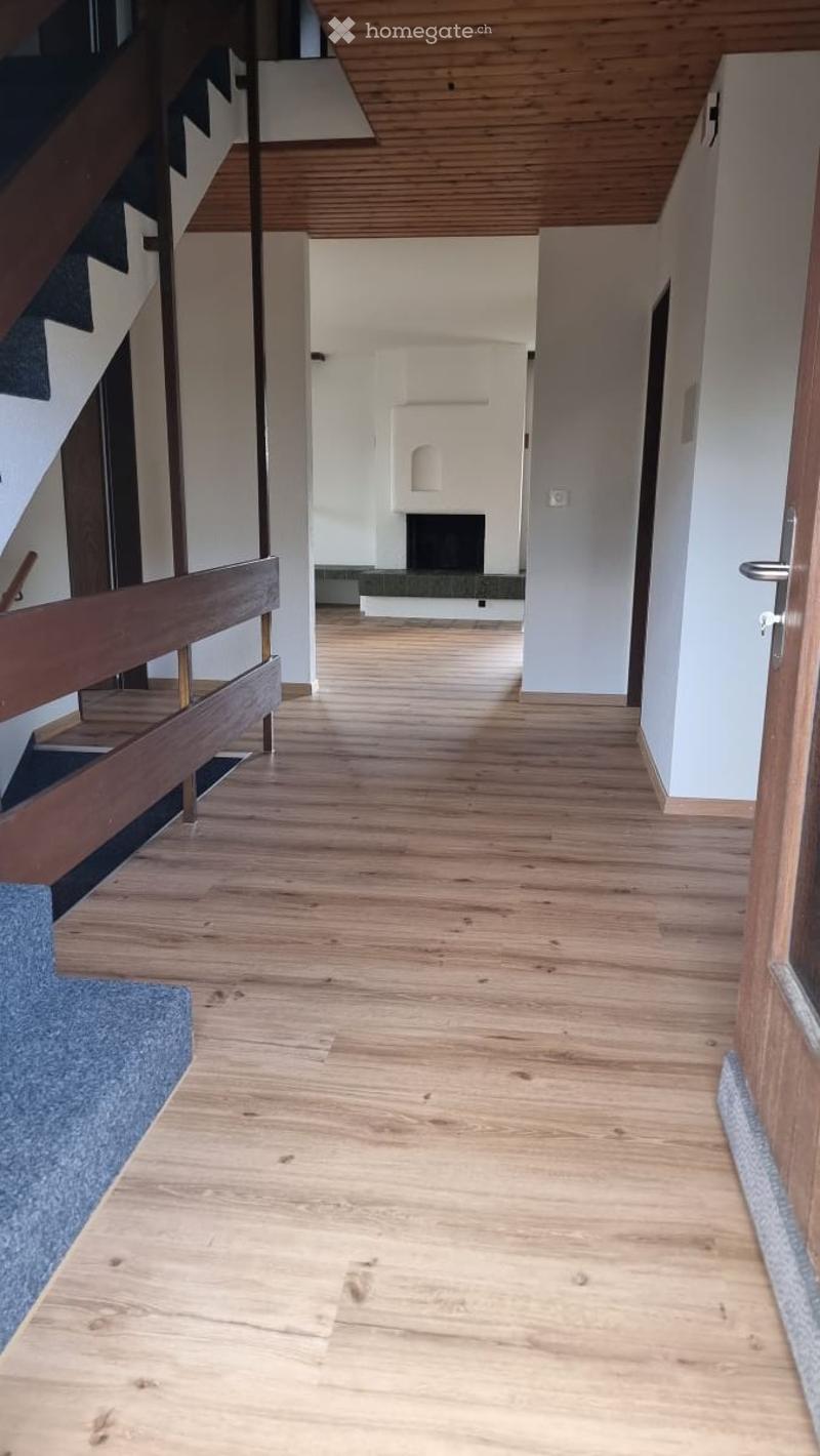 Bright hallway with wooden flooring, white walls, and a staircase leading upwards. A fireplace is visible in the background.
